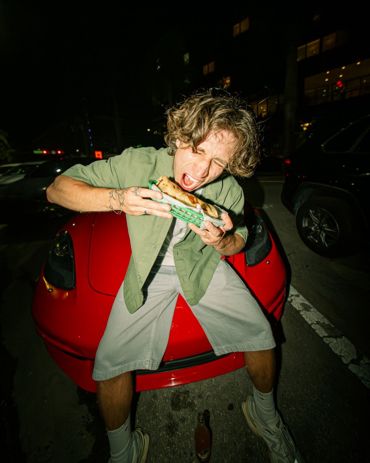 Sergio Teran, founder of WHATEVR Creative Studios, biting into a burrito on the hood of a red sports car at night