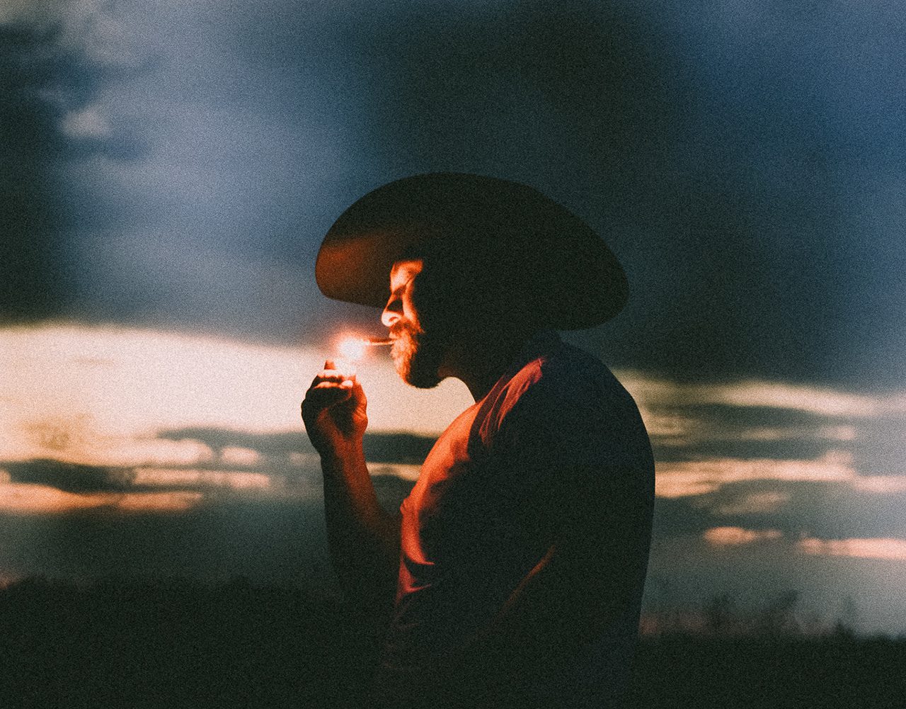 Man in a cowboy hat lighting a cigarette against a dusk sky, REDBILLY campaign